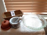 Photo showing all items including loaf pan with tag, marble mortar and pestle, glass pie plate, and terracotta garlic baking dish on table near window with blinds