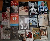 Top-down photo showing 16 Canadian fiction and non-fiction books arranged in a grid on a wooden surface. Two Rick Mercer books are prominently displayed with visible titles. Other authors include Alice Munro, Frances Itani, Fred Sasakamoose, Robertson Davies, Michael Ondaatje, and Christy Ann Conlin.