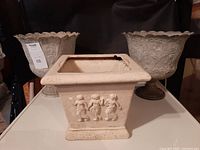 Photo showing two matching ornate metal flower pots with scalloped edges and one square plaster flower pot with relief decoration of children.