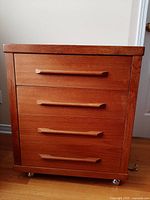Front view of the teak chest of drawers with four drawers and horizontal handles on caster wheels.