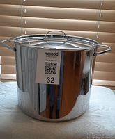 Front and side view of the stainless steel stock pot with lid on a white tablecloth in front of wooden blinds.