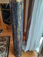 Tall cylindrical floor vase with iridescent crackle black and gold finish, standing on floor next to curtains and fireplace, showing its full height and textured surface.