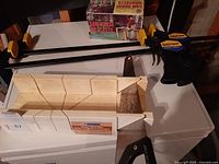 Photo showing two large Quick-Grip bar clamps next to a white plastic mitre saw box and a hand saw resting on a white surface with saw horse bracket box in background.
