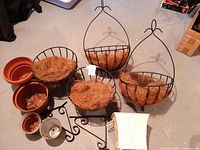 Photo showing two wrought iron hanging baskets lined with coconut fiber matting, one smaller (12") and one larger (14") without base, along with other gardening pots and items around.