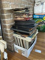 Stack of vinyl albums and 45 RPM records piled on the floor showing original sleeves and some loose covers, accompanied by boxes containing more vinyl.