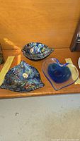 Three art glass dishes on a wooden surface: two colorful leaf-shaped dishes with Venetian art glass labels and one blue and clear heart-shaped dish.