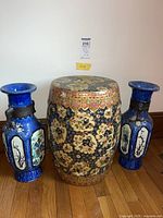 Front view of Asian garden stool centered between two blue artisan vases on wooden floor against white wall.