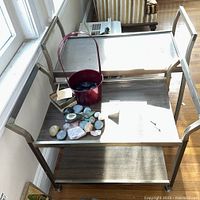 Two metal framed rolling TV carts with veneer shelves, one larger and one smaller, placed near a window with items on shelves for scale and reference.