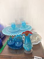 Fenton blue opalescent hobnail cake stand, two matching hobnail candleholders on the stand, with additional blue hobnail dishes, decorative blue-red rimmed art glass pitcher, and white hobnail glass dish on wooden surface.
