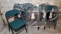 Eight folding chairs, greenish teal fabric upholstery with patterned design, stacked and wrapped in plastic.