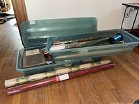 Three large translucent blue plastic storage bins with lids containing assorted rolls of festive wrapping paper and ribbon roll, and some gift bags.