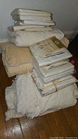 Stack of vintage bedspreads folded: cream colored woven fabric with fringe, beige chenille or textured bedspread, bundle of packaged sheets in plastic wrapping.