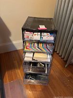 Front view of black plastic five drawer organizer with transparent drawers containing office supplies such as binder clips, fasteners, Post-it notes and floppy disks.