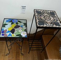 Two plant stands next to each other; one with floral butterfly stained glass tile top and decorative wrought iron base, the other with geometric tile top on taller wrought iron base with slatted bottom shelf.