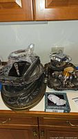 Wide view of stacked silver plated trays, glassware items, and boxed items on countertop.