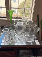 Overview photo showing variety of glass and crystal vases on a table by window, including green fluted vase and multiple clear shapes.