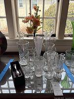 Photo showing a collection of 10 clear glass and crystal vases of different shapes and sizes arranged on a glass-topped table with some floral stems inside one vase.
