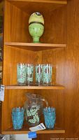 Clear glass pitcher with green leaf decoration and multiple glasses with blue pattern on wooden shelf