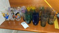 The full lot of 27 assorted vintage glasses spread on a wooden surface. Includes clear glasses with cartoon prints, blue and green tinted glasses, and floral decorated glasses.