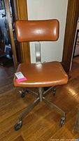 Front angled view of vintage Domore office chair showing brown vinyl seat, backrest, metal base with caster wheels on hardwood floor.