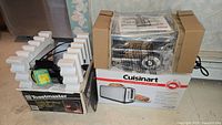 Two boxed appliances: Toastmaster Pizzelle/Waffle Baker in box with styrofoam, and Cuisinart Artisan Bread Toaster boxed with visible packaging and manual on top.