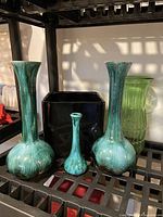 Photo showing three turquoise glass vases of varying sizes and shapes and a green ribbed glass vase behind them on a shelf.
