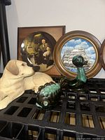 Two green glazed ceramic animal figurines (duck and hedgehog) with beige ceramic dog figurine, displayed in front of a Norman Rockwell decorative plate featuring an elderly man and children.