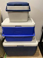 Three stacked Coleman coolers: small white and blue cooler on top, medium blue cooler in the middle, and large blue cooler on bottom.
