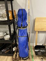 Blue golf bag with zippered compartments containing assorted golf clubs; adjacent to a chair and shelving unit with kitchenware.