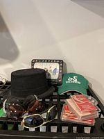 Photo showing black felt top hat, green Lochness baseball cap, NHL hockey game board, two pairs of sunglasses on a black crate with decks of playing cards