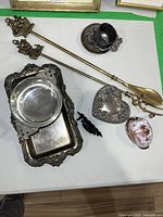 Metal trays and decorative items arranged on white surface including ornate trays, heart-shaped dish, candle snuffer, and decorative egg