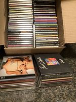 Box filled with stacked music CDs showing some album cover titles in front and multiple CDs underneath in jewel cases.