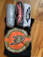 Four Stanley Cup Champion t-shirts, one visible black t-shirt with Anaheim Ducks logo and three others tightly rolled in plastic packaging, colors grey, red, white, on wooden floor.