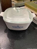 White square ceramic casserole dish with blue floral design and clear glass lid on black surface