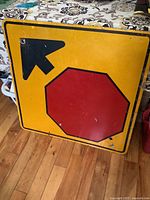 Front side of the large metal traffic sign showing a yellow background with a red octagonal stop symbol and a black arrow pointing diagonally left and upwards.