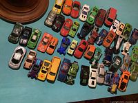 Multiple older diecast toy cars including various colors, makes, models, and styles displayed on a turquoise surface, some showing surface wear and paint chips.