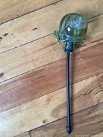 Full view of green glass snail garden stake with metal rod on wooden floor.