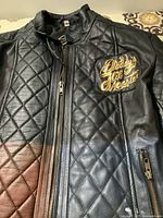 Front view showing black quilted leather jacket with zipper and gold lettering 'Daddy's Lil Monster' on chest.