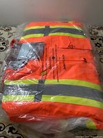 Packed high visibility orange work jacket with fluorescent yellow and silver reflective stripes, folded inside a sealed plastic bag on a patterned surface.