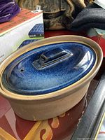 Stoneware crock with natural brown base and blue glazed lid with handle and vent hole, placed on wooden surface among other items.