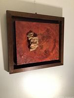 Framed mixed media artwork with a textured central form on a red background, hung on wall.
