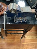 Side view of the small blue bench vice sitting on a black wooden table showing the metal jaws, handle, and rotating clamp base.