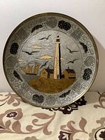 Front view of brass plate showing lighthouse, sailing ship, birds, and houses with decorative border