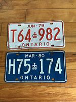 Photo showing two Ontario license plates: white with red letters T64 982 dated June 1979, and blue with white letters H75 174 dated March 1980, placed on wooden floor.