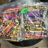 Two sealed plastic bags each filled with various Crayola crayons of multiple colors shown on a round black surface.