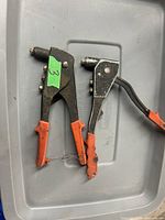Two hand rivet tools resting on a grey plastic surface, showing orange plastic-coated handles and metal jaws for rivet application.