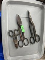 Photo showing three used metal cutting snips with metal handles and blades, placed inside a gray plastic tray, showing signs of rust and wear.
