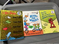 Three vintage children's books: One Fish Two Fish Red Fish Blue Fish Party Edition by Dr Seuss, Bears on Wheels by Stan and Jan Berenstain, and Can You Tell Me How to Get to Sesame Street featuring Elmo held on a container.