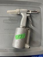 Side view of a silver metal air rivet tool with pistol grip and trigger, lying on a gray surface. Green label marked '11' is visible.