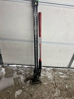Full view of the car bumper jack showing perforated black steel bar, red handle, and base plate on a concrete floor with snow nearby.